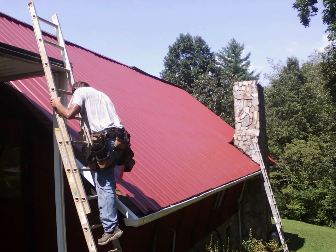 Roofer on a ladder beside a red metal roof and stone chimney — Soffit & Fascia Repair by Roofing Brothers in Kingsford Heights